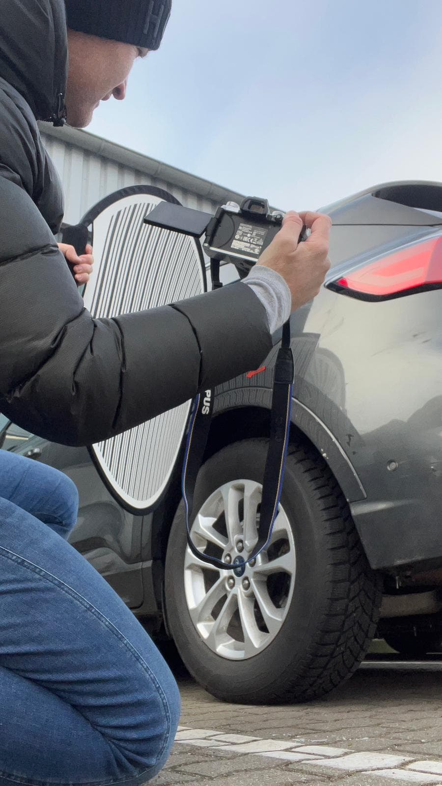 Sebastian Paul fotografiert mit Kamera und Reflektorleuchte die Heckpartie eines grauen SUV bei einer Begutachtung im Freien
